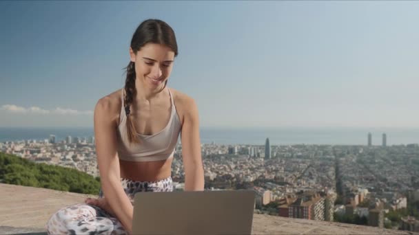 Kvinna som sitter i lotus poserar med laptop utomhus. Flicka skriva på bärbar dator i staden — Stockvideo