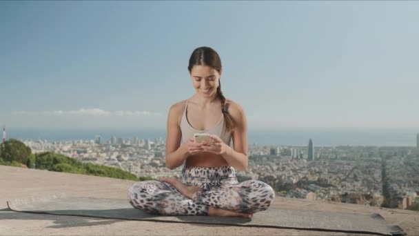 Žena kouká na obrazovku chytrého telefonu ve městě. Dáma sedící v lotosu pózuje na podložce — Stock video