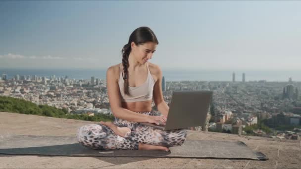 Chica usando portátil en la ciudad de Barcelona. Mujer de yoga sentada en la pose de loto en la estera — Vídeo de stock