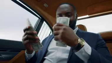 Yakından Afrikalı bir adam arabada dolar kokusu alıyor. Araçta paralı bir adam oturuyor.