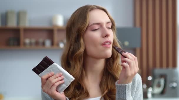 Close up jovem mulher comendo chocolate na cozinha. Menina relaxada desfrutando de chocolate — Vídeo de Stock