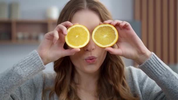 Молодая женщина держит два кусочка апельсина на кухне. Девушка корчит рожи апельсинами — стоковое видео