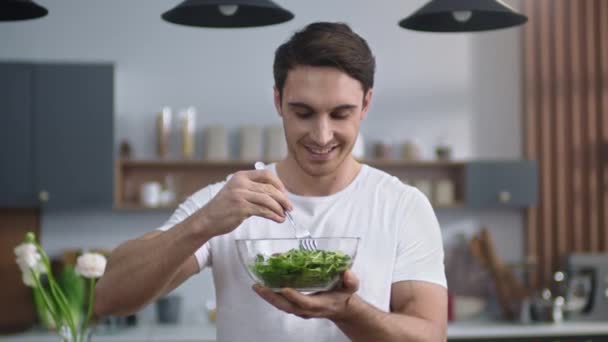 Χαμογελαστή άνθρωπος τρώει φρέσκια σαλάτα στην κουζίνα του σπιτιού. Αθλητικός τύπος που απολαμβάνει υγιεινά τρόφιμα — Αρχείο Βίντεο