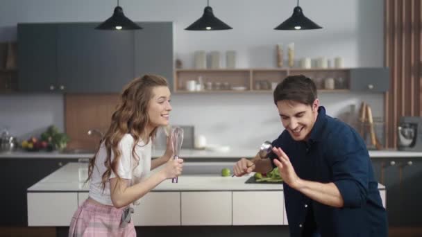 Casal alegre cantando música em utensílios de cozinha em casa cozinha. Amigos se divertindo — Vídeo de Stock