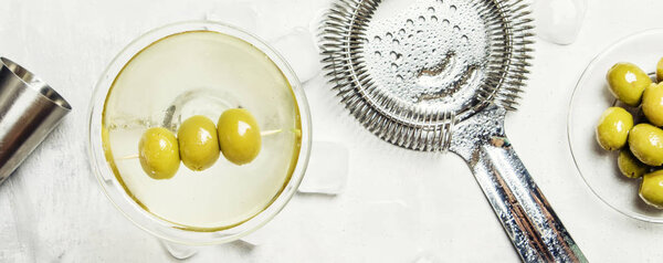Classic alcoholic cocktail with dry vermouth and green olives, bar tools, gray background, top view