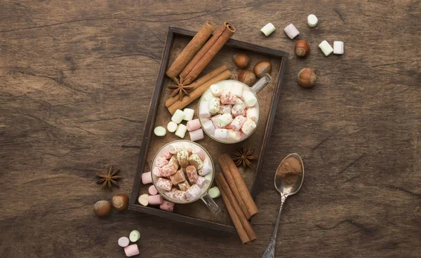 Hot winter or autumn drink with cocoa, white chocolate, spices and colorful marshmallows in cups on vintage wooden background with knitted warm sweater, top view with copy space