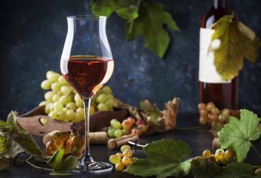 Şarap tatma konseptinde pembe şarap kadehi. Üzüm ve yapraklarla dolu gri bir arkaplan. Boşluğu kopyala