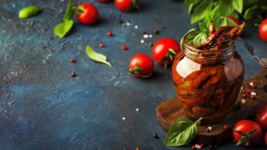 Italian Sun Dried tomatoes in olive oil with green basil and spices in glass jar on blue kitchen table, copy space clipart