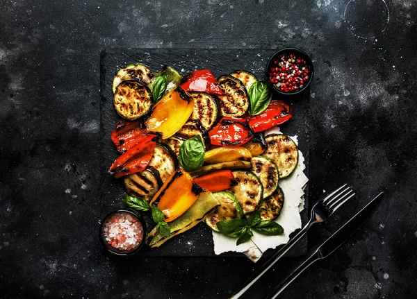 Grilled multicolored vegetables, aubergines, zucchini, pepper with green basil on serving stone board on black background, top view