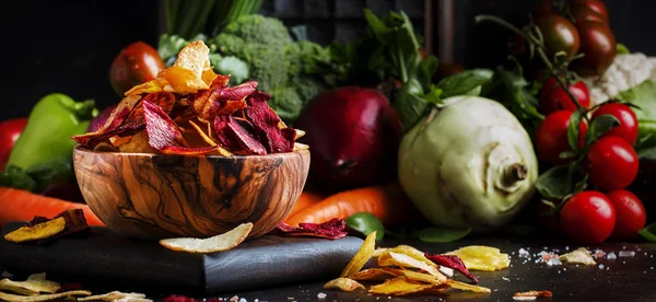 Vegan snacks, multicolored vegetable chips in wooden bowl, set of fresh farmer vegetables, still life