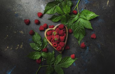 Fresh raspberries in bowl in shape heart on dark background, top view
