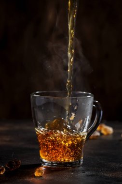 Kahverengi arka planda doğal buharla cam fincana siyah çay dökülüyor.