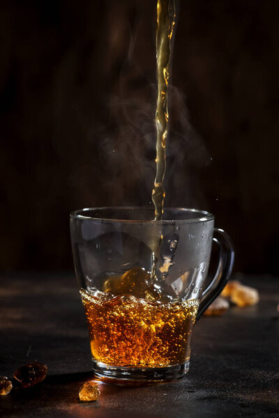 Black tea pouring into glass cup with natural steam on brown background