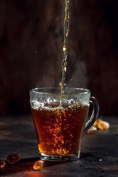 Black tea pouring into glass cup with natural steam on brown background