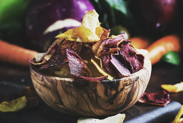 Vegan snacks, multicolored vegetable chips in wooden bowl and set of fresh farmer vegetables, rustic still life, selective focus