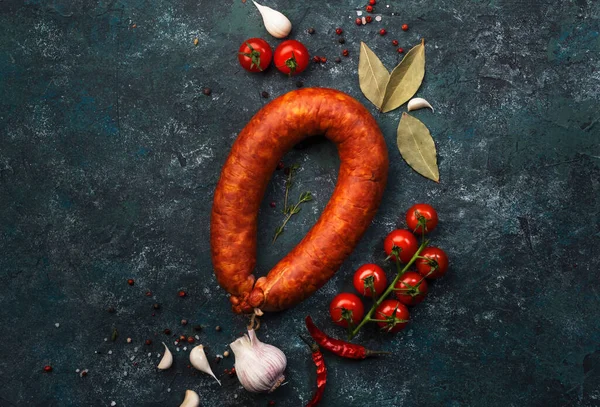 Smoked sausage on grey kitchen table background with aromatic herbs and spices, tomatoes and garlic, natural organic farm meat product, top view, copy space