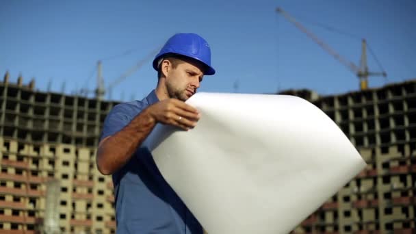 Architect in helmet on building background. Engineer at a construction site looking drawing.  Man is holding a drawing next to the house under construction. Modern building, multi-storey building. — Stock Video