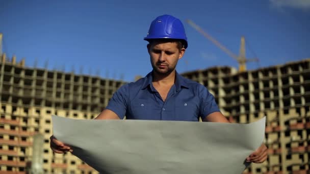 工程师在看图纸的施工现场。建筑师在头盔上建设背景。男人拿着一个绘图旁边在建房屋。现代建筑，多层大厦. — 图库视频影像
