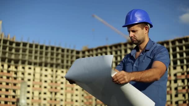 Engenheiro em um canteiro de obras olhando desenho. Arquiteto em capacete no fundo do edifício. Edifício moderno, edifício de vários andares. Homem está segurando um desenho ao lado da casa em construção . — Vídeo de Stock