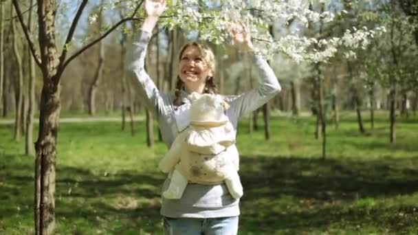 スリングで赤ちゃんを持つ若い母親。ママは、公園で子供と一緒に歩いています。スプリングパーク、ママがスリングで赤ちゃんを運ぶ. — ストック動画