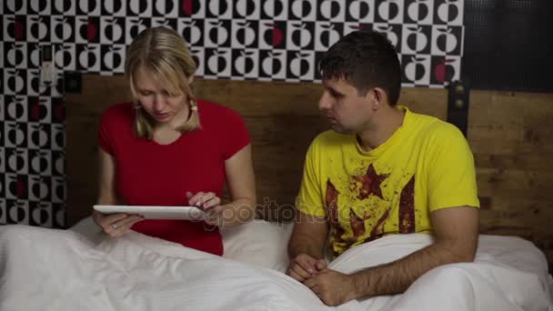 En kvinna med en tablett dator, och mannen är arg bredvid honom. Ett gift par på sängen, Internetberoende. — Stockvideo