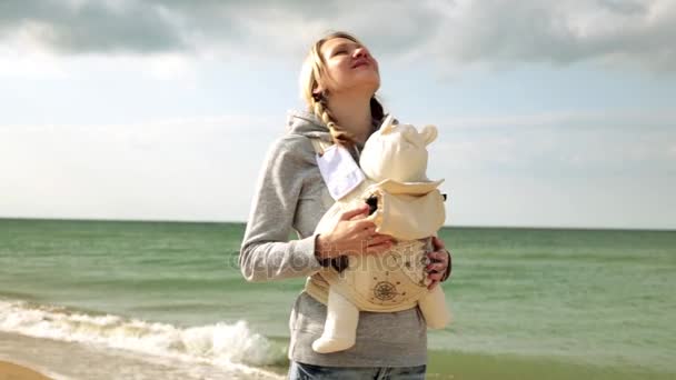 Mamma med barn på stranden. En kvinna med ett barn vid havet. Kvinna med baby i sele på naturen. Mamma med ett barn i en sele. — Stockvideo