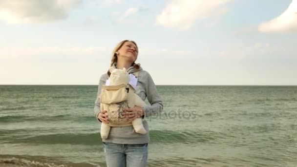 Mom with a baby in a sling. A woman with a child by the sea. Mother with baby on the beach. — Stock Video