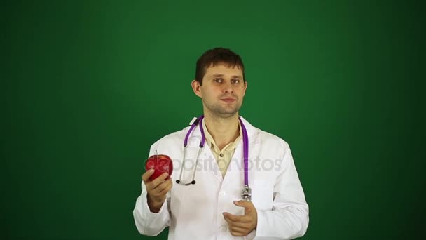 En manlig läkare är att äta ett äpple. Tandläkare biter ett äpple. Läkare på en grön bakgrund som innehar ett äpple. — Stockvideo