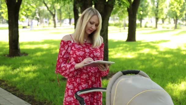 Una chica con una tableta camina con su bebé. Mamá y su hijo usan una tableta. Mujer con cochecito y tablet . — Vídeos de Stock