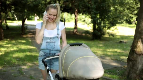 Uma mulher com um carrinho de bebé está a falar ao telefone. Mãe caminha com a criança e telefones . — Vídeo de Stock