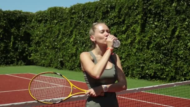 Sportliche Frau mit Plastikflasche nach dem Tennistraining. Mädchen auf dem Tennisplatz trinkt Wasser. — Stockvideo