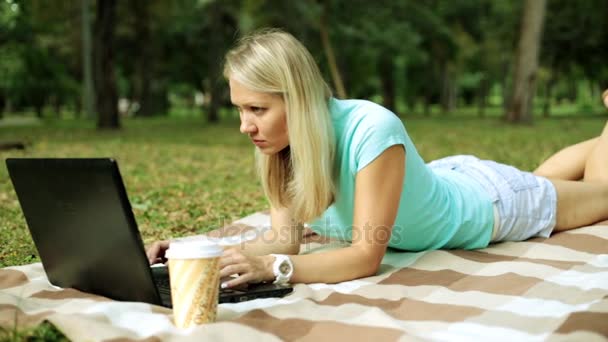 Jonge vrouw met laptop in het park. Het meisje is liggend op een deken met een laptop. — Stockvideo