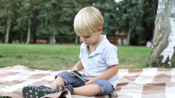 Een kind speelt met een smartphone in het park op het gras. — Stockvideo
