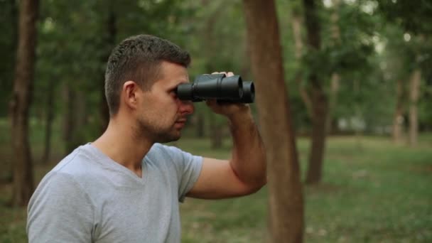 Un tipo con prismáticos contra los árboles. Un joven mira a través de los prismáticos . — Vídeos de Stock