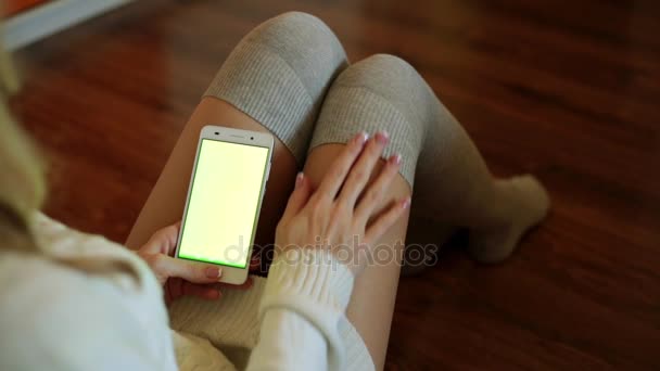 Una hermosa mujer se sienta en una habitación y sostiene un teléfono inteligente con una pantalla verde . — Vídeos de Stock