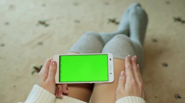 Bir kadın yere oturur ve yeşil bir ekran ile bir akıllı telefon tutar.