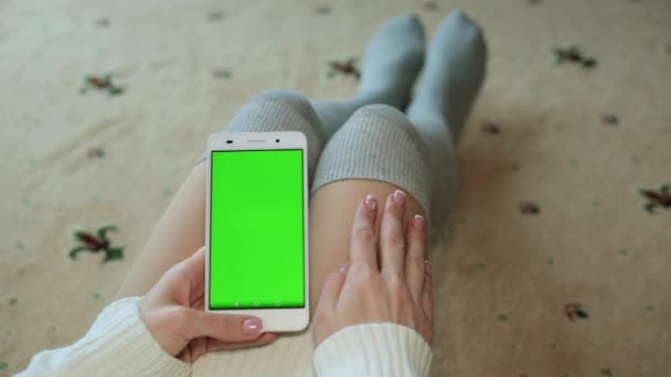 一个性感的女人坐在地板上, 并持有一个智能手机与绿色屏幕为您的内容. — 图库视频影像