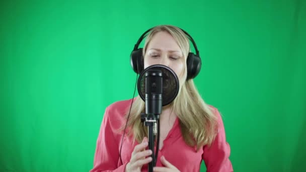 Uma mulher em fones de ouvido canta em um microfone em um fundo verde . — Vídeo de Stock