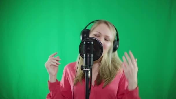 Yeşil bir arka plan üzerinde bir mikrofona kulaklık genç bir kadın şarkı söylüyor. — Stok video