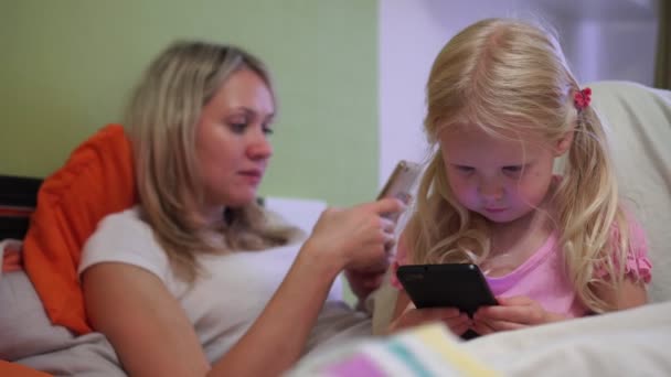 Girl child looks in a smartphone, in the background mom uses a smartphone. — Stock Video