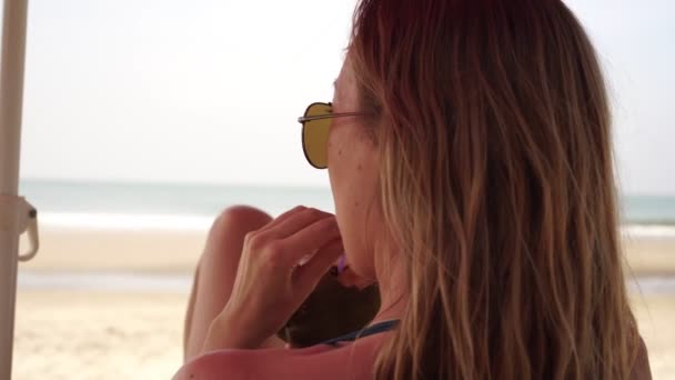 En ung kvinna kopplar av vid havet, dricker kokos och solar på stranden. — Stockvideo