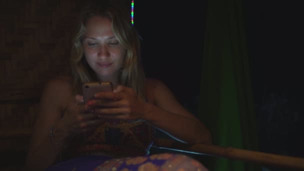 白いブロンドの女性は暗闇の中で夜にスマートフォンにメッセージを書きます — ストック動画