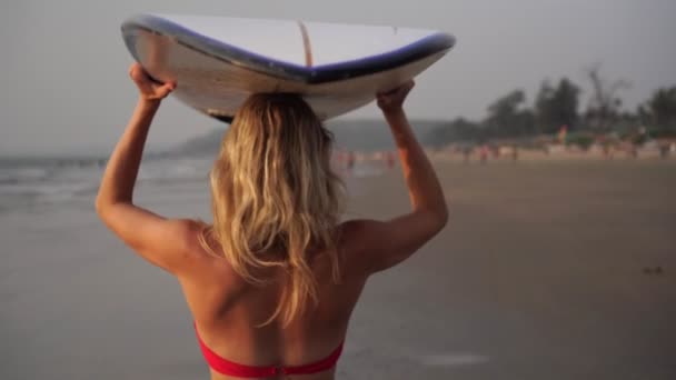 Mooie jonge vrouw in een rood badpak loopt langs het strand met een surfplank — Stockvideo