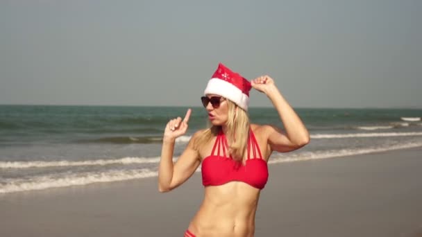 En ung kvinna i baddräkt dansar på en havsstrand i en nyårshatt. — Stockvideo