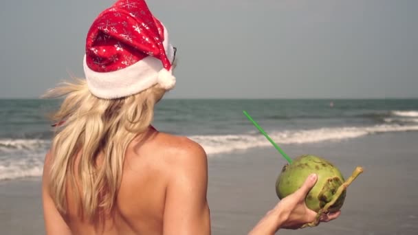 Gyönyörű nő télapó kalapban kókuszt iszik a tengerparton. Karácsony a tengeren — Stock videók