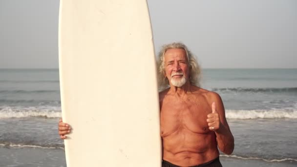 Velho homem maduro com prancha de surf na praia mostrando polegares para cima — Vídeo de Stock