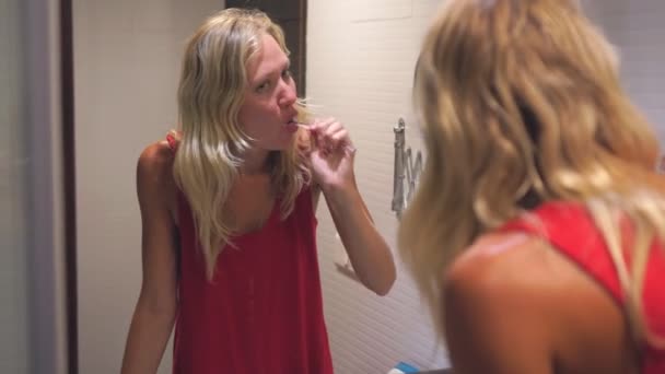 Joven mujer atractiva rubia se cepilla los dientes en el baño delante del espejo — Vídeo de stock