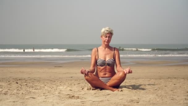 Una donna matura pratica yoga o meditazione sulla spiaggia. Nonna nella posizione del loto — Video Stock