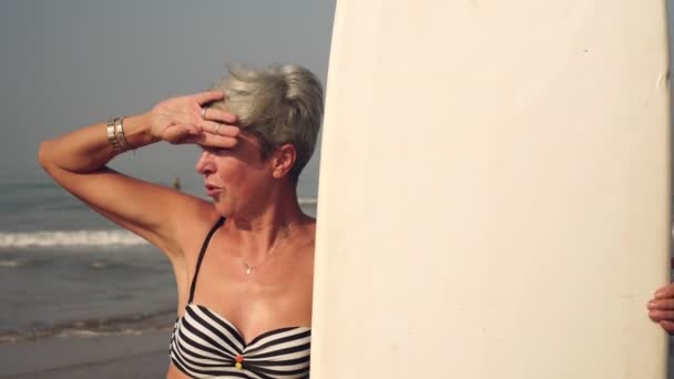 Mujer madura en la playa con una tabla de surf. Abuela con un oleaje en el fondo del mar — Vídeo de stock