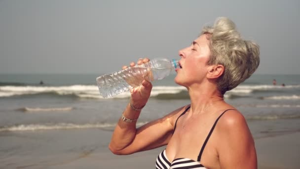 Rijpere aantrekkelijke vrouw drinkt water uit een fles op het strand — Stockvideo